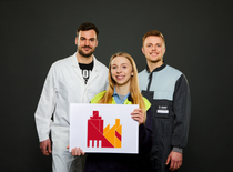 3 Azubis in Arbeitskleidung halten das Key Visual der BASF Würzburg